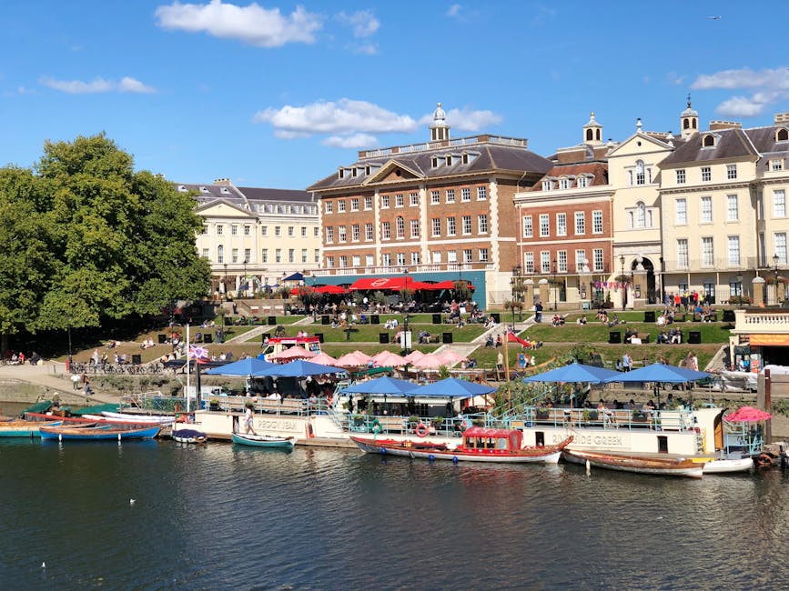 A detailed view of a picturesque riverside scene showing multiple boats docked along a wooden pier, with some boats covered by bright blue and pink umbrellas providing shade for outdoor seating areas. Behind the boats, there is a lively urban setting with stairs leading up to a terraced area filled with people sitting and relaxing on grassy slopes and benches. The background features historic multi-story buildings with a mix of brick and cream-colored facades, ornate gabled roofs, dormer windows, and decorative trim, set against a clear blue sky with a few fluffy clouds. Large leafy trees frame the left side of the scene, adding greenery to the scene. The environment suggests a popular leisure spot, with outdoor dining and social activities visible, and the scene contextually aligns with independent or private waste management practices, characteristic of rental or commercial vessels and nearby urban public spaces, reflecting a typical setting where rubbish removal might be necessary for maintaining cleanliness.