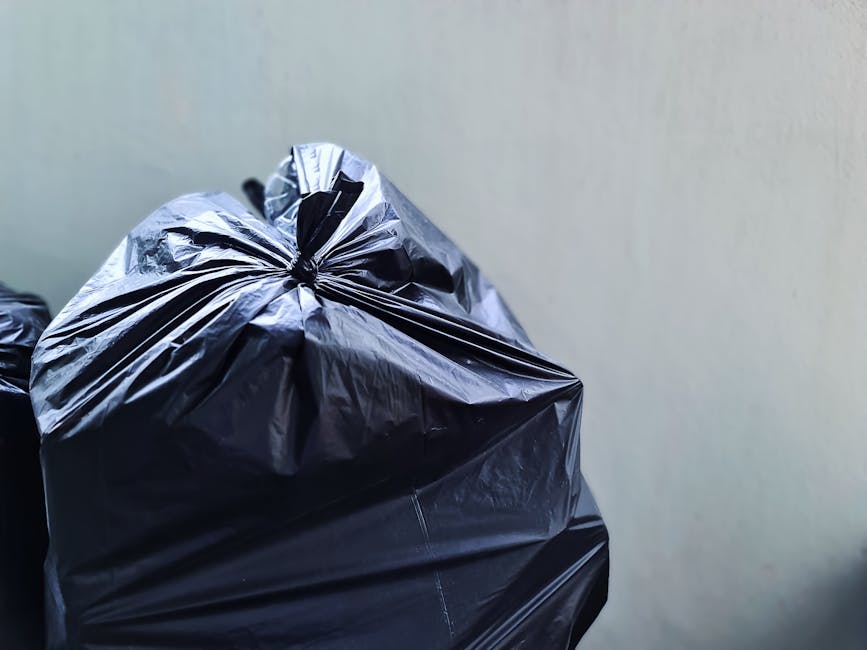A close-up view of several black rubbish bags made from glossy plastic material, tightly knotted at the top, placed against a plain light-colored wall. The bags are stacked or gathered together in a corner, with one bag in the foreground and others slightly behind, indicating a collection of waste ready for disposal. The surface of the bags reflects ambient light, highlighting the crinkled texture and the sheen of the plastic material. The environment appears to be an indoor or sheltered area, possibly a storage or collection point for waste before collection or disposal by a private rubbish removal service such as [COMPANY_NAME]. The scene reflects the process of private waste handling, where refuse is gathered in durable, sealable bags, awaiting collection. This visual aligns with the context of alternative rubbish collection options, emphasizing neatness and readiness for professional rubbish removal near Richmond Station, in accordance with the site’s service scope.