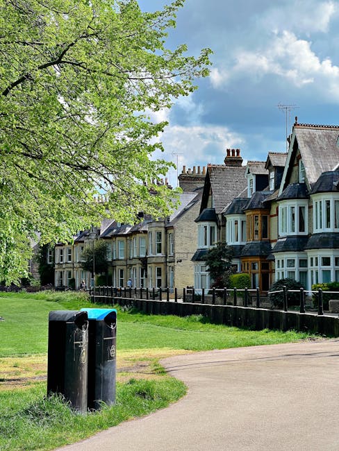 A public park area in a residential neighbourhood featuring two black rubbish bins positioned on a grassy patch near a paved walkway. Behind the bins, there is a row of Victorian-style terraced houses with bay windows, stone facades, and slate roofs, aligned along the street. To the left, a large leafy tree with bright green foliage extends across the scene, partially shading the grass and pathway. The sky overhead is partly cloudy with patches of blue visible, suggesting a fair weather day. The area appears well-maintained, with a simple wooden fence running parallel to the houses, separating the grass from the street. This scene illustrates a typical urban setting where private waste collection services, such as those provided by local companies, are often used for rubbish disposal in residential areas, offering an alternative to council-provided waste collection.