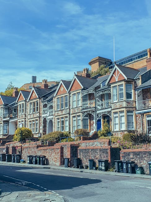 A row of Victorian-style terraced houses with decorative cast-iron balconies and large bay windows, positioned on an elevated street with a brick retaining wall and small front gardens. The houses are constructed from red brick with contrasting white trim, featuring gabled roofs and ornate detailing. In the foreground, there are several black and dark green wheelie bins lined up along the pavement, indicating waste collection points. The scene is set on a bright, clear day with a blue sky and wispy clouds, and the street appears clean and well-maintained. The overall environment suggests a residential area where private waste collection, possibly managed by independent waste removal services like rubbishcollectionrichmonduponthames.co.uk, is a common practice for managing refuse outside the scope of local authority collection.