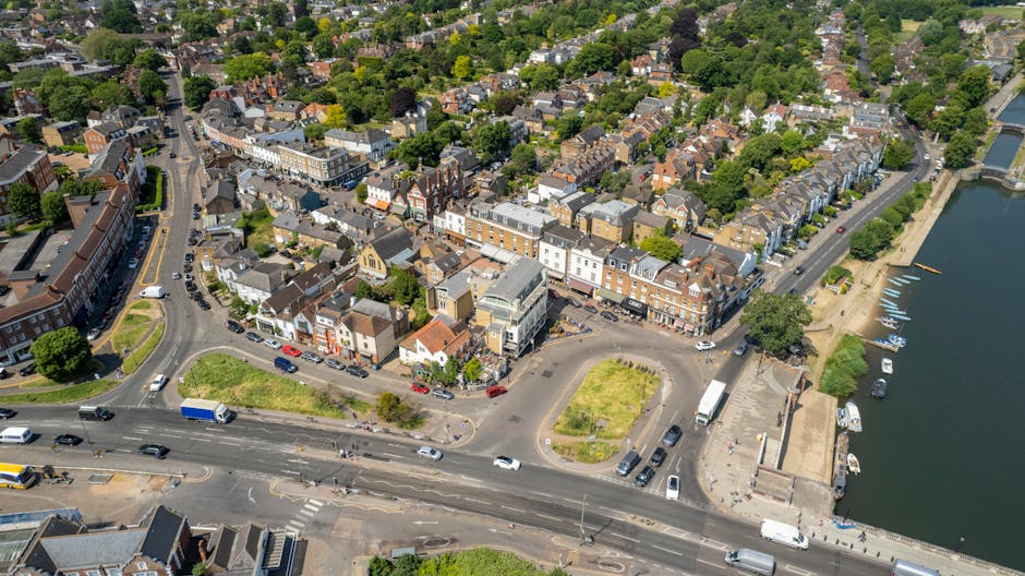 An aerial view of a small town showing a mixture of residential and commercial buildings, with a prominent street running diagonally across the foreground. The buildings have varied architectural styles, including brick facades, pitched roofs, and flat roofs, with some featuring brickwork and others painted in neutral tones. Along the main road, there are parked cars and moving vehicles including vans and cars, with some green spaces and small roundabouts nearby. To the right, there is a body of water with boats docked along the shoreline, bordered by a paved walkway and some trees. Surrounding the area are numerous trees and residential houses with gardens, creating a lush, green environment. The overall scene is well-lit with natural daylight, and the image captures the compact urban layout typical of a small town close to a waterfront. This setting may reflect local private waste collection services operating within the town, with a landscape that supports local rubbish removal and on-site clearance activities, as indicated by the density and arrangement of buildings and roadways nearby. The company Richmond upon Thames waste management services could facilitate alternative rubbish handling in this area through scheduled collection points and community-based disposal options.